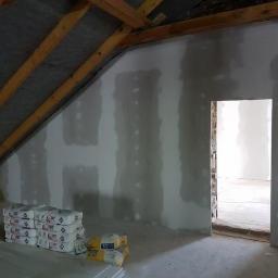 RiW Remonty i Wykończenia - Poddasze z ukośnym sufitem, ściany pokryte płytami kartonowo-gipsowymi, widoczne łączenia wypełnione masą szpachlową, w tle otwór drzwiowy, na podłodze ułożone płyty i worki z zaprawą.