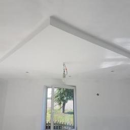 RiW Remonty i Wykończenia - Wnętrze pokoju po szpachlowaniu ścian i sufitu, widoczne ślady po łatach na ścianach, otwarte okno z widokiem na zieleń, surowy żyrandol na kablu.