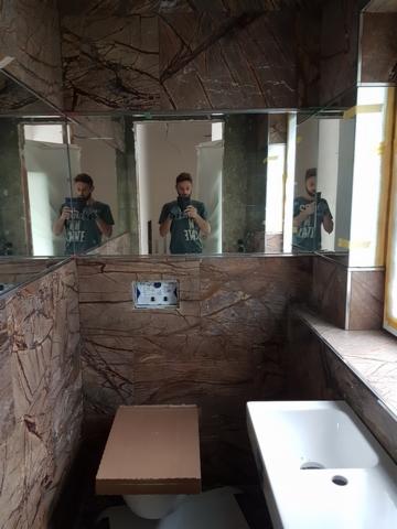 Wykończona łazienka z brązowymi płytkami imitującymi marmur na ścianach, lustra w wąskich ramach na górze, biała umywalka z prostokątnym zlewem, toaleta z deską zabezpieczoną kartonem, widoczny...