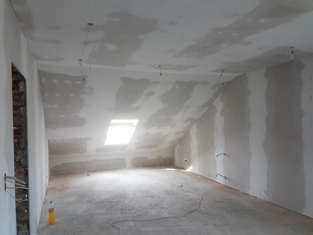 Poddasze w trakcie remontu: ściany i sufit pokryte wstępną warstwą szpachli, widoczne kable elektryczne wystające ze ścian, okno dachowe w skosie, butelka z napojem na podłodze.