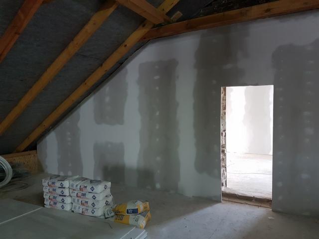 Poddasze z ukośnym sufitem, ściany pokryte płytami kartonowo-gipsowymi, widoczne łączenia wypełnione masą szpachlową, w tle otwór drzwiowy, na podłodze ułożone płyty i worki z zaprawą.