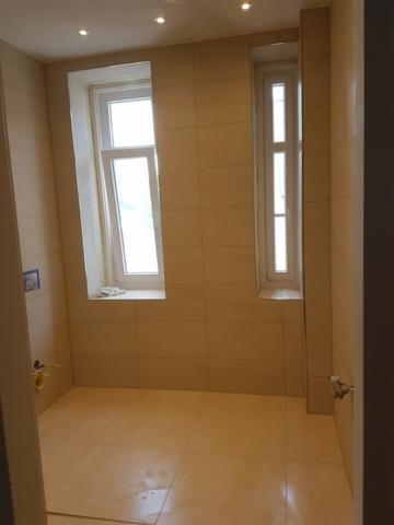 Jasna łazienka z beżowymi, kwadratowymi płytkami na ścianach i podłodze, dwa okna o różnej wysokości, widoczne ślady po montażu armatury.