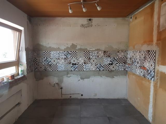 Wnętrze kuchni w trakcie remontu: częściowo ułożone płytki dekoracyjne na ścianie, surowy stan ścian, szara podłoga z płytek, widoczne instalacje wodne i elektryczne, okno z widocznym parapetem.