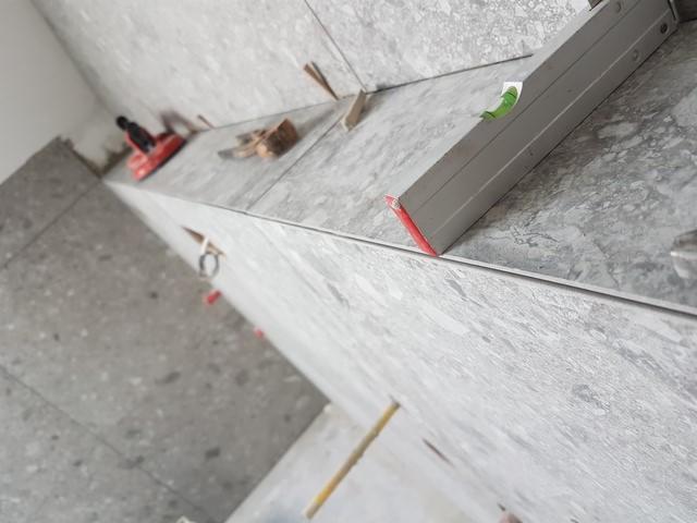 Poziomica spoczywająca na świeżo ułożonych, szarych płytkach o nieregularnym wzorze, widoczne narzędzia glazurnicze i kliny dystansowe.