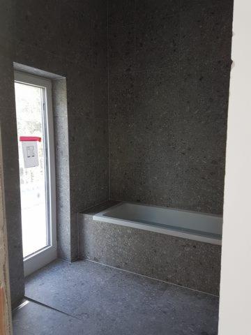 Wykończona łazienka z szarymi płytkami imitującymi kamień na ścianach i podłodze, widoczna wanna obudowana tymi samymi płytkami oraz częściowo przesłonięte drzwi balkonowe.