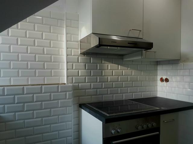 Wykończona kuchnia z białymi, błyszczącymi płytkami typu metro na ścianie, czarnym blatem kuchennym i srebrnym okapem Bosch. Widoczne gniazdka elektryczne bez osprzętu.