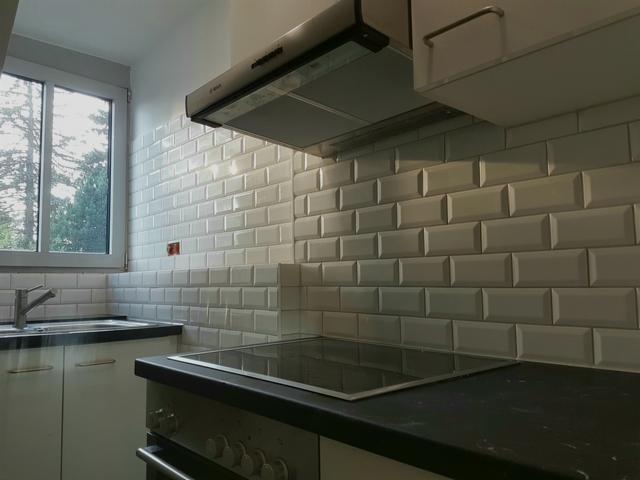 Kuchnia z białymi, wypukłymi płytkami typu metro, czarnym blatem i płytą indukcyjną, widok na okno z drzewami w tle.