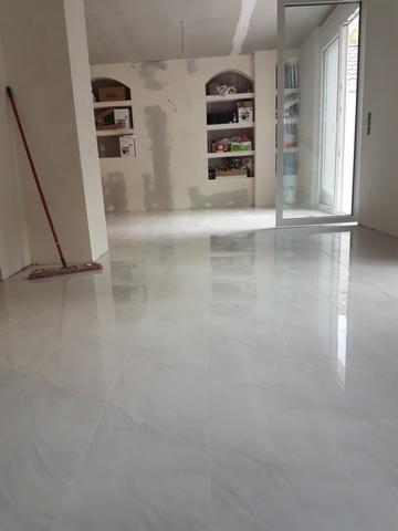 Błyszcząca, jasnoszara podłoga z płytek imitujących marmur w nowym pomieszczeniu, widoczne wnęki ścienne z półkami, ślady prac wykończeniowych na ścianach i mop oparty o ścianę.