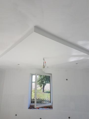 Wnętrze pokoju po szpachlowaniu ścian i sufitu, widoczne ślady po łatach na ścianach, otwarte okno z widokiem na zieleń, surowy żyrandol na kablu.