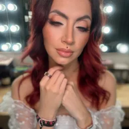 Portret kobiety z rudymi włosami, w delikatnym makijażu z błyszczącymi ustami i subtelnym cieniem na powiekach, pozującej na tle lustra z żarówkami