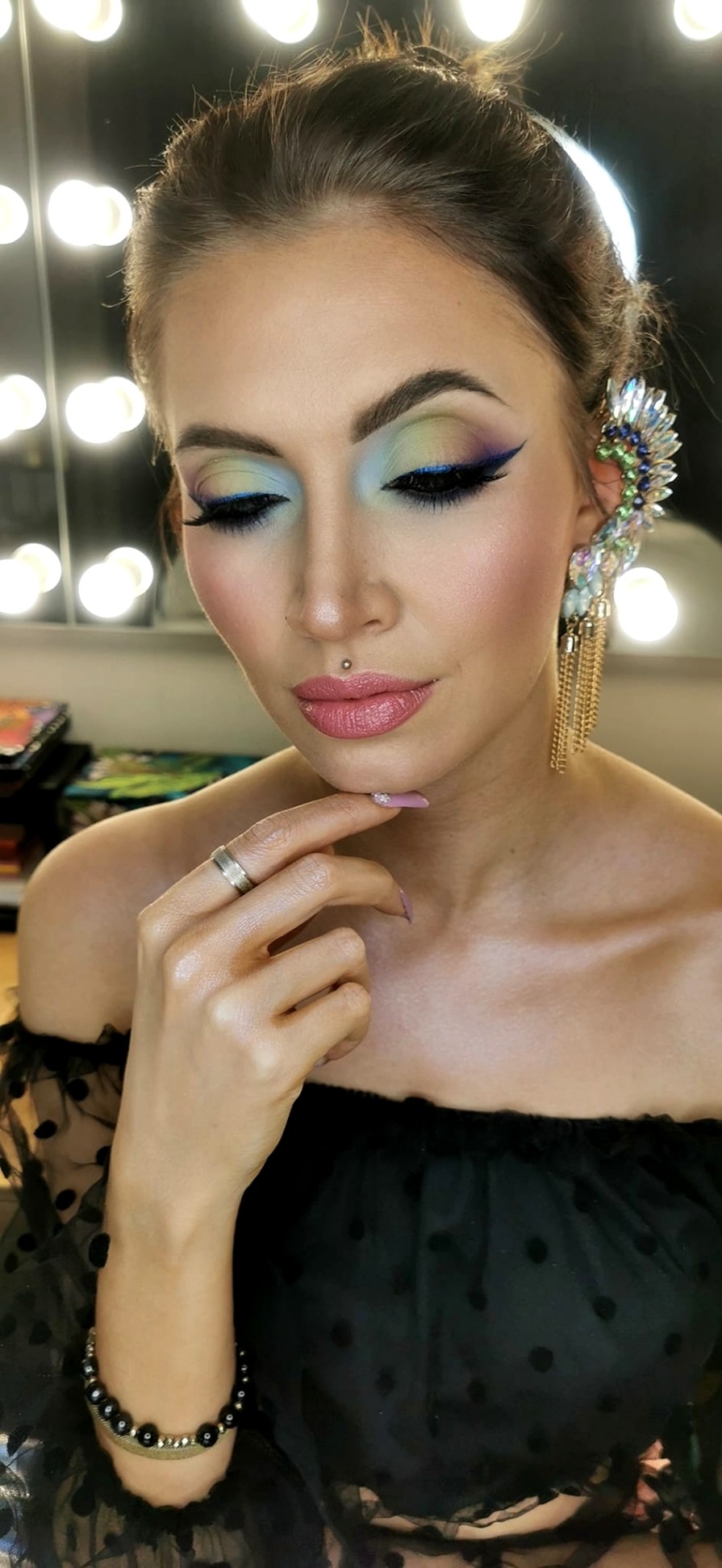 Portret kobiety z profesjonalnym makijażem: tęczowe cienie, czarny eyeliner, błyszczące usta, subtelny kolczyk w wardze i ozdobny nausznik z łańcuszkami.