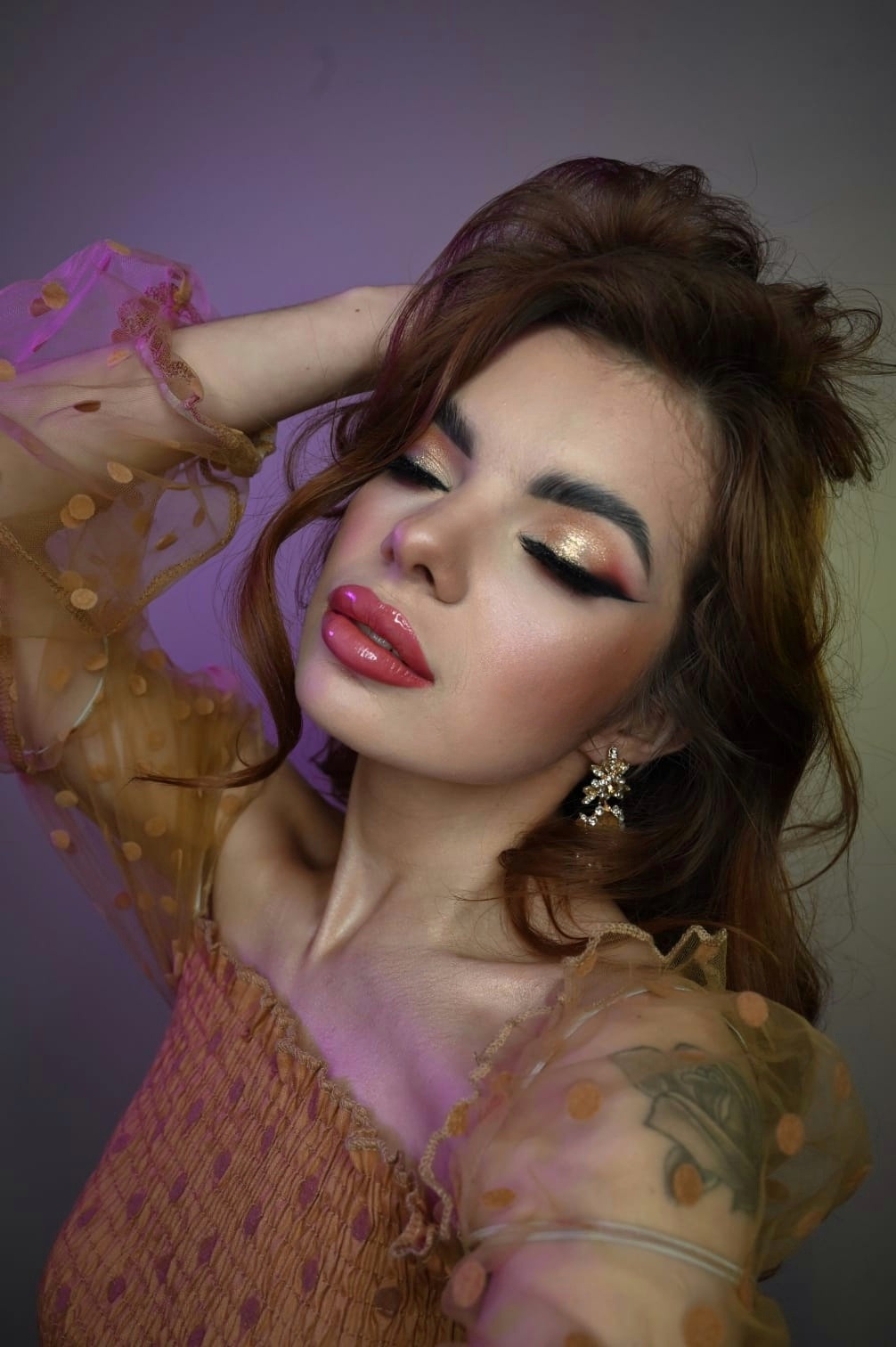 Portret kobiety z zamkniętymi oczami, prezentującej profesjonalny makijaż: błyszczące złote cienie, wyraźna kreska, mocno podkreślone brwi i błyszczące, powiększone usta. Uwagę przyciągają kolczyki...