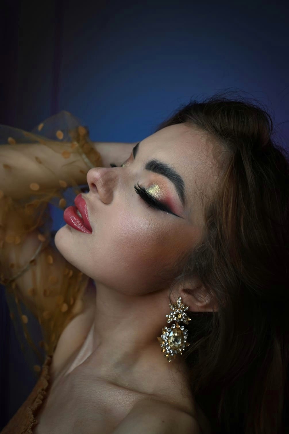 Profil kobiety z zamkniętymi oczami, prezentującej złoto-różowy makijaż oczu z czarną kreską, błyszczące usta i bogato zdobione kolczyki na tle niebiesko-fioletowym.