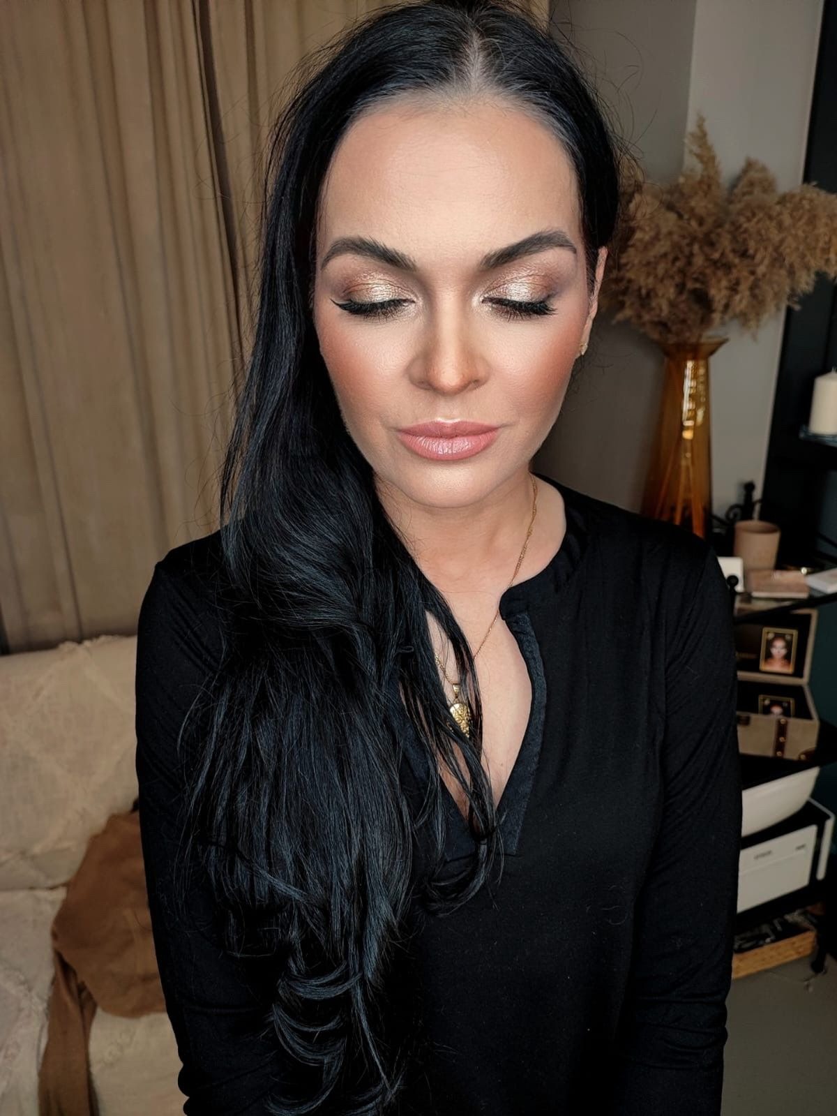 Portret kobiety z zamkniętymi oczami, prezentujący złoty makijaż oczu z delikatną kreską, podkreślone kości policzkowe i naturalne usta, czarne długie włosy opadające na czarną bluzkę, w tle beżowa...