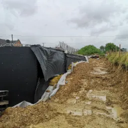 Wykop pod fundamenty z zabezpieczoną ścianą wykopu, widoczne worki z piaskiem i folia ochronna, błotniste podłoże po deszczu.