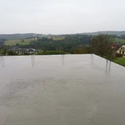 Zalana betonem płyta fundamentowa z wystającymi prętami zbrojeniowymi, na tle pagórkowatego krajobrazu z zabudowaniami w pochmurny dzień.