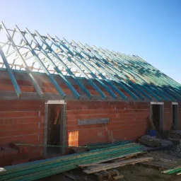 Surowy stan domu w trakcie budowy: ściany z czerwonej cegły, drewniana konstrukcja dachu pomalowana na niebiesko, widoczne materiały budowlane i rusztowania.