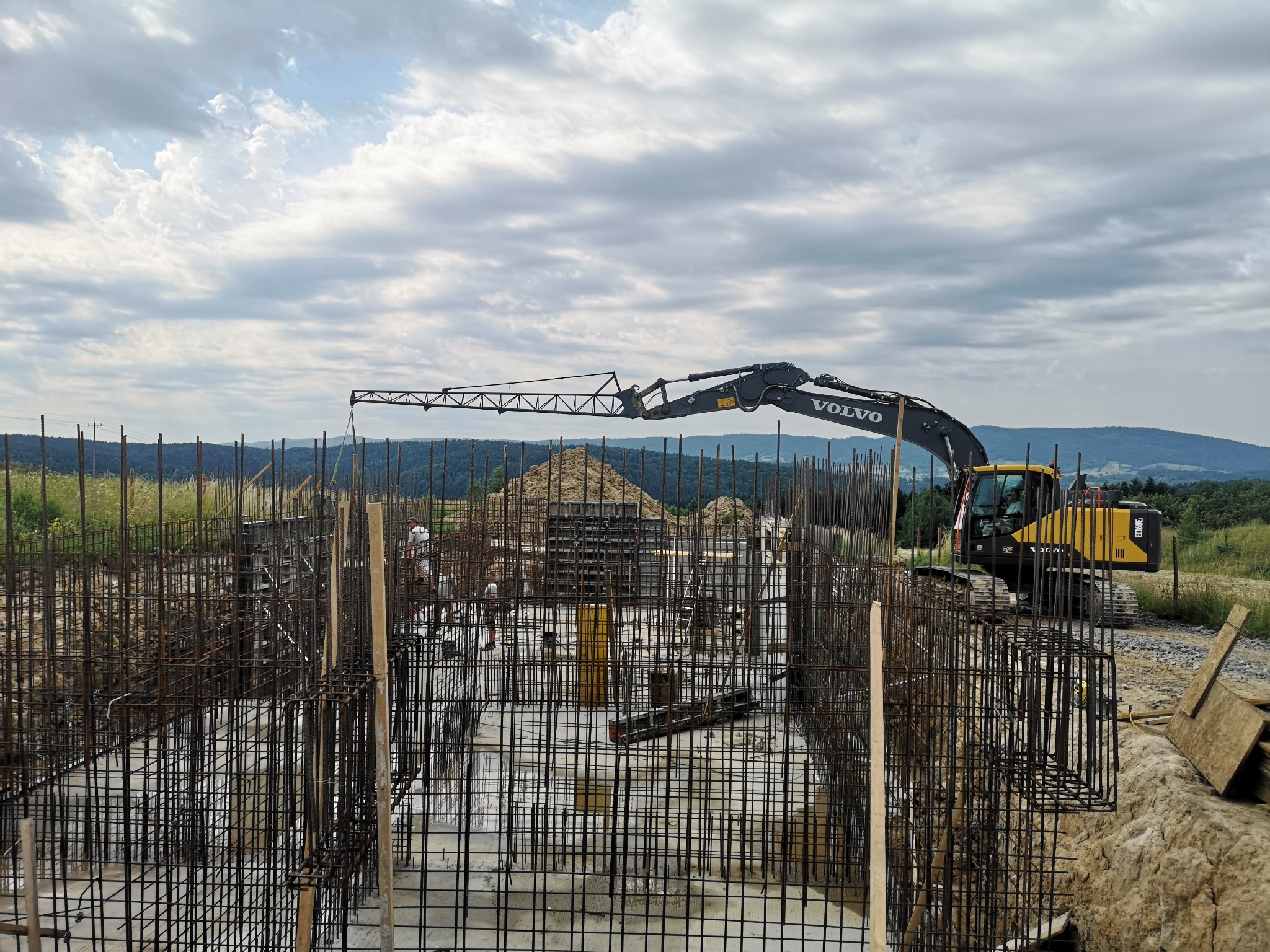 Zbrojenie fundamentów z widoczną koparką Volvo w tle, krajobraz zalesionych wzgórz pod pochmurnym niebem.