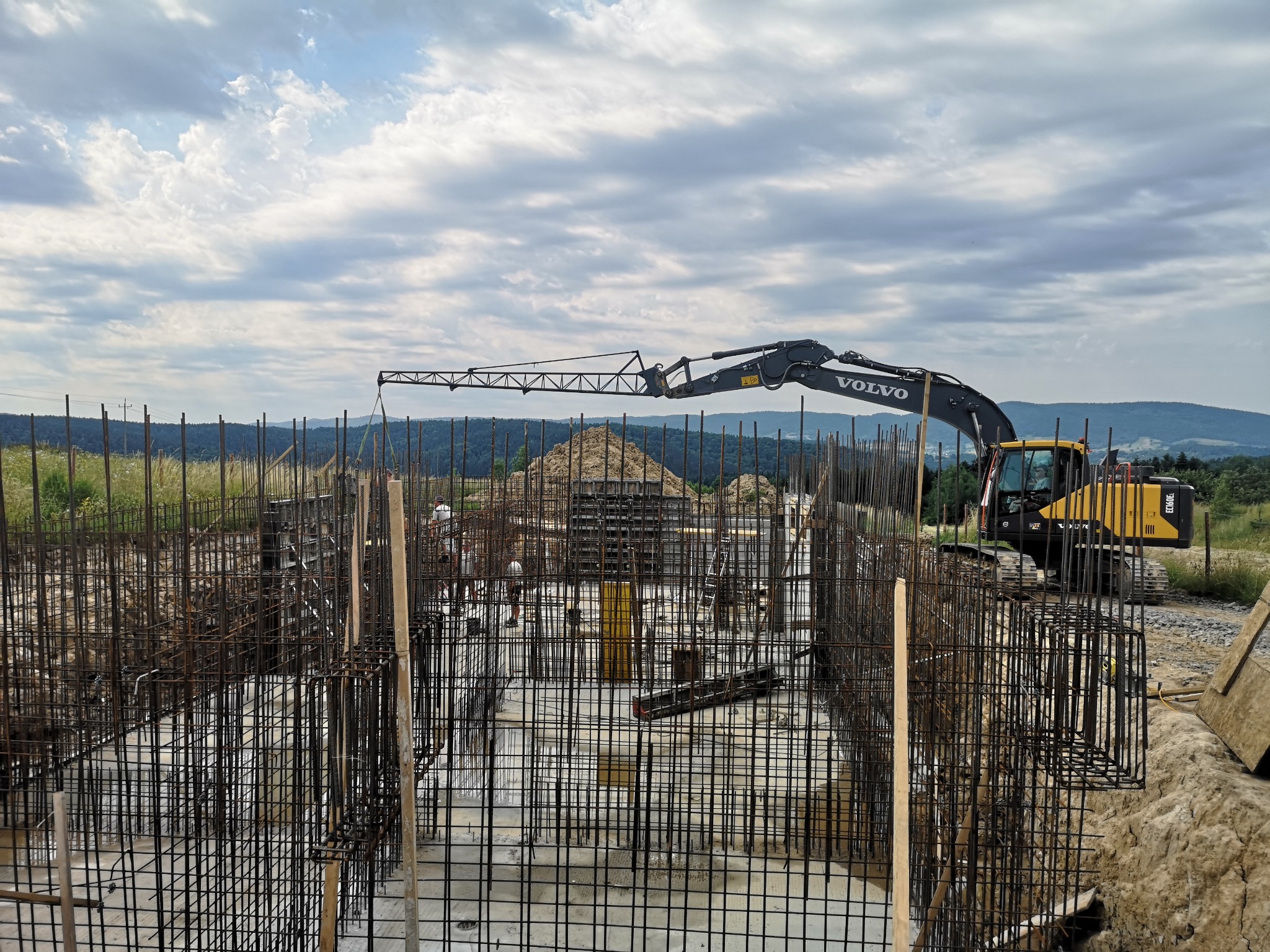 Siatka zbrojeniowa fundamentów w trakcie budowy, widoczny ciężki sprzęt Volvo i kopiec ziemi w tle, krajobraz zalesionych wzgórz pod pochmurnym niebem.