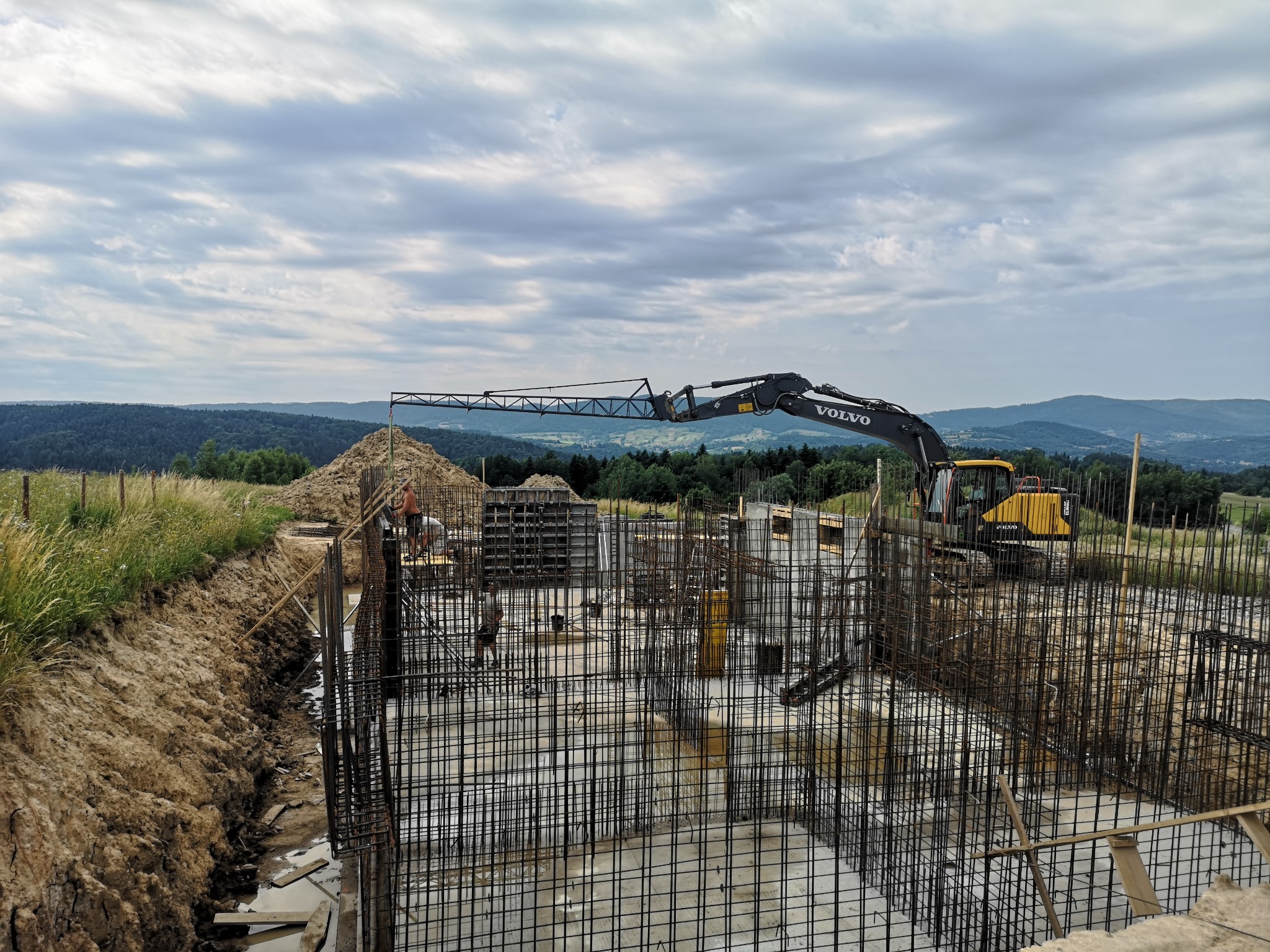 Zbrojenie fundamentu z widocznym betonem, szalunkami i częścią koparki Volvo na tle pagórkowatego krajobrazu pod pochmurnym niebem.