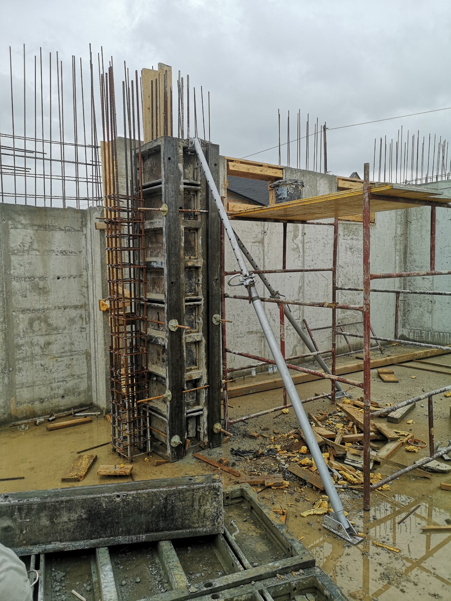 Szare betonowe ściany fundamentowe z wystającą stalową siatką zbrojeniową, drewniane szalunki podtrzymywane metalowymi stemplami, widoczne rusztowanie i błoto na placu budowy w pochmurny dzień.