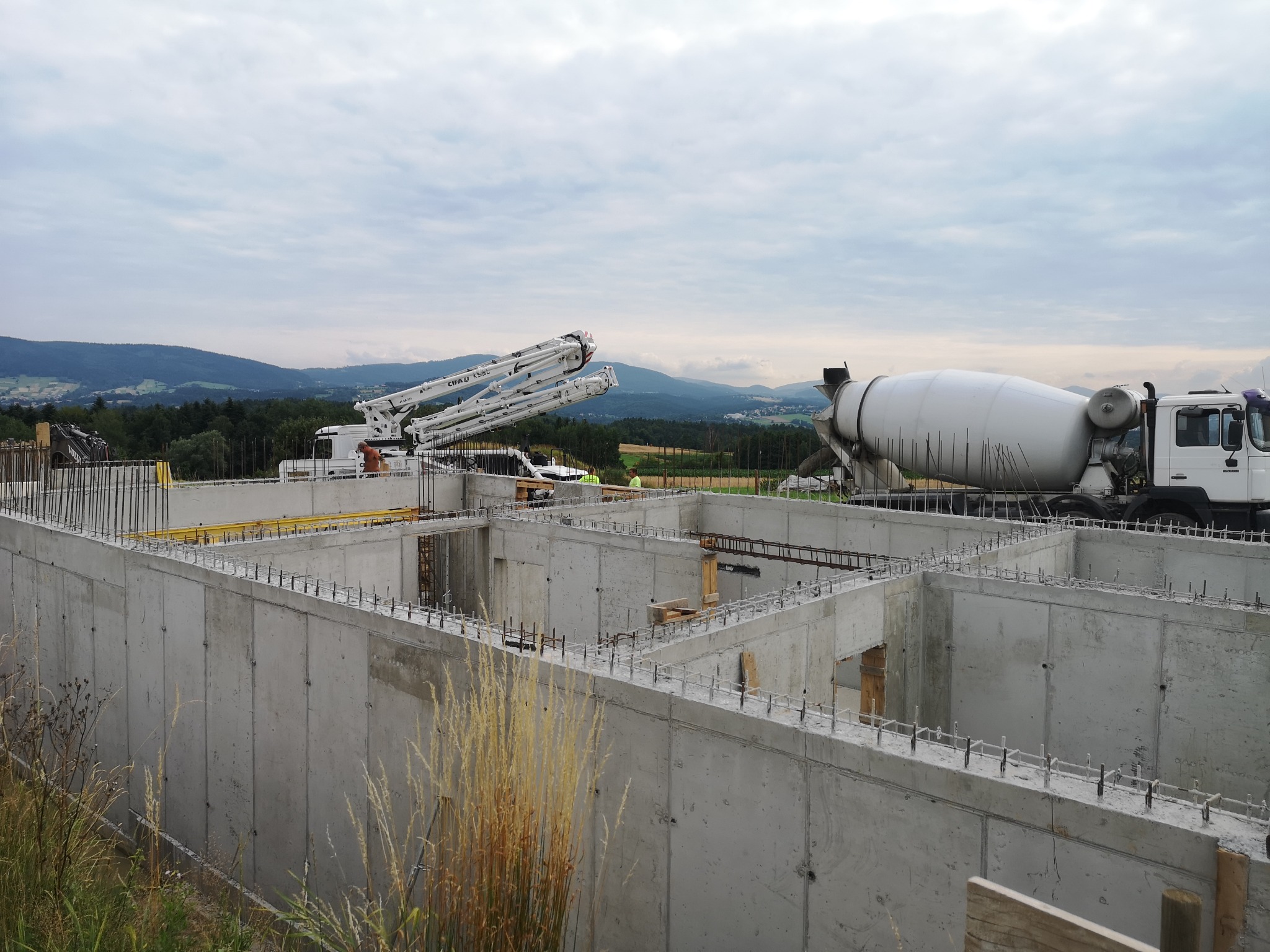 Wylany betonowy fundament domu z wystającymi prętami zbrojeniowymi, widoczna betoniarka i pompa do betonu na tle pagórkowatego krajobrazu pod pochmurnym niebem.