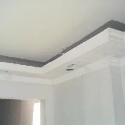 Wnętrze w trakcie wykańczania: sufit z ozdobnym gzymsem i przygotowanymi otworami na punktowe źródła światła, ściany w jasnym kolorze, widoczne kable.