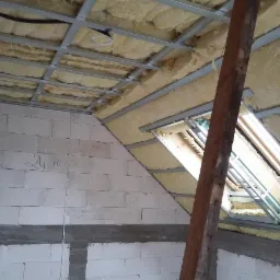 Poddasze w trakcie ocieplania wełną mineralną, widoczne metalowe profile sufitowe, okno dachowe i fragment ściany z bloczków z betonu komórkowego z napisem '24 Kotek'.
