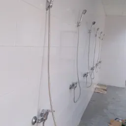 Rząd zamontowanych chromowanych baterii prysznicowych z wężami i słuchawkami na białej, kafelkowanej ścianie w szatni lub natryskach publicznych, widoczne narzędzia i karton na podłodze.