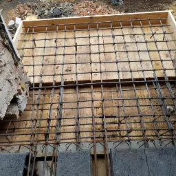 Zbrojenie płyty fundamentowej z drewnianym szalunkiem, widok z góry na plac budowy z fragmentami ceglanego muru.