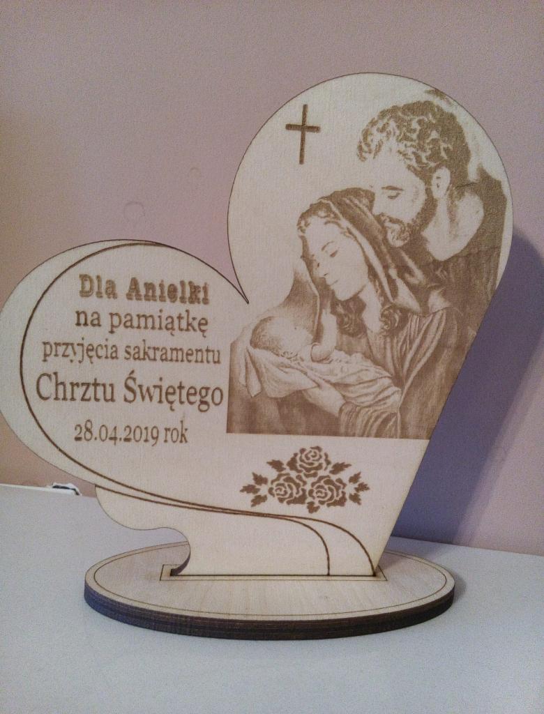 Drewniana pamiątka Chrztu Świętego w kształcie serca z wygrawerowanym wizerunkiem Świętej Rodziny i napisem 'Dla Aniołki na pamiątkę przyjęcia sakramentu Chrztu Świętego 28.04.2019 rok', ozdobiona...