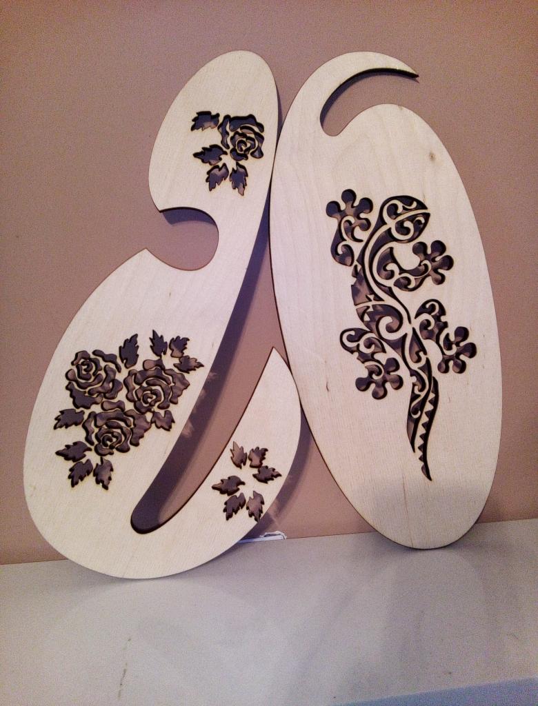 Dwa drewniane, laserowo wycinane elementy dekoracyjne: jeden w kształcie litery 'J' z motywem róż, drugi owalny z wyciętą sylwetką jaszczurki.