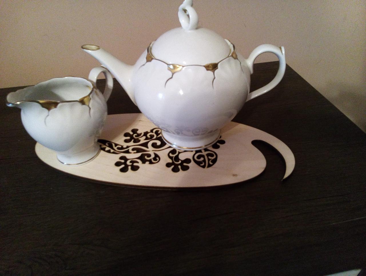 Biały serwis do herbaty z dzbankiem i mlecznikiem, zdobiony złotymi akcentami, na jasnej, drewnianej tacy z wyciętym wzorem.