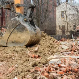 Łyżka koparki zanurzona w gruzie i ziemi po rozbiórce budynku, z fragmentami ceglanej ściany w tle.