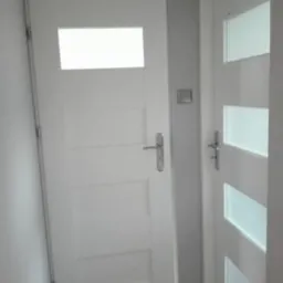 Dwie białe, nowoczesne drzwi wewnętrzne z przeszkleniami, jedna z pojedynczym, prostokątnym oknem u góry i czterema okrągłymi otworami na dole, druga z trzema poziomymi pasami szkła.