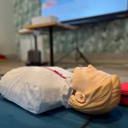 Manekin Little Anne do nauki pierwszej pomocy, leżący na niebieskiej macie, w tle ekran z prezentacją i stolik.