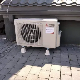 Zewnętrzna jednostka klimatyzacji Mitsubishi Electric zamontowana na białych podstawkach na szarej kostce brukowej, widoczny budynek z szarą elewacją i rynną.