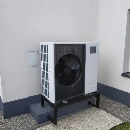 Biała pompa ciepła powietrze-woda na czarnym metalowym stelażu, zamontowana przy jasnej ścianie budynku, otoczona drobnym żwirem.