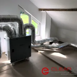 Szary rekuperator z podłączonymi, izolowanymi przewodami wentylacyjnymi w pomieszczeniu na poddaszu z oknem i widokiem na zewnątrz.