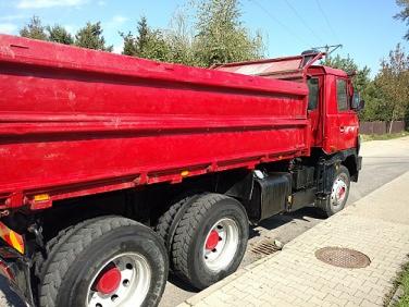 Czerwona wywrotka zaparkowana na poboczu, widok z boku, z widocznymi oponami, skrzynią ładunkową i elementami podwozia.