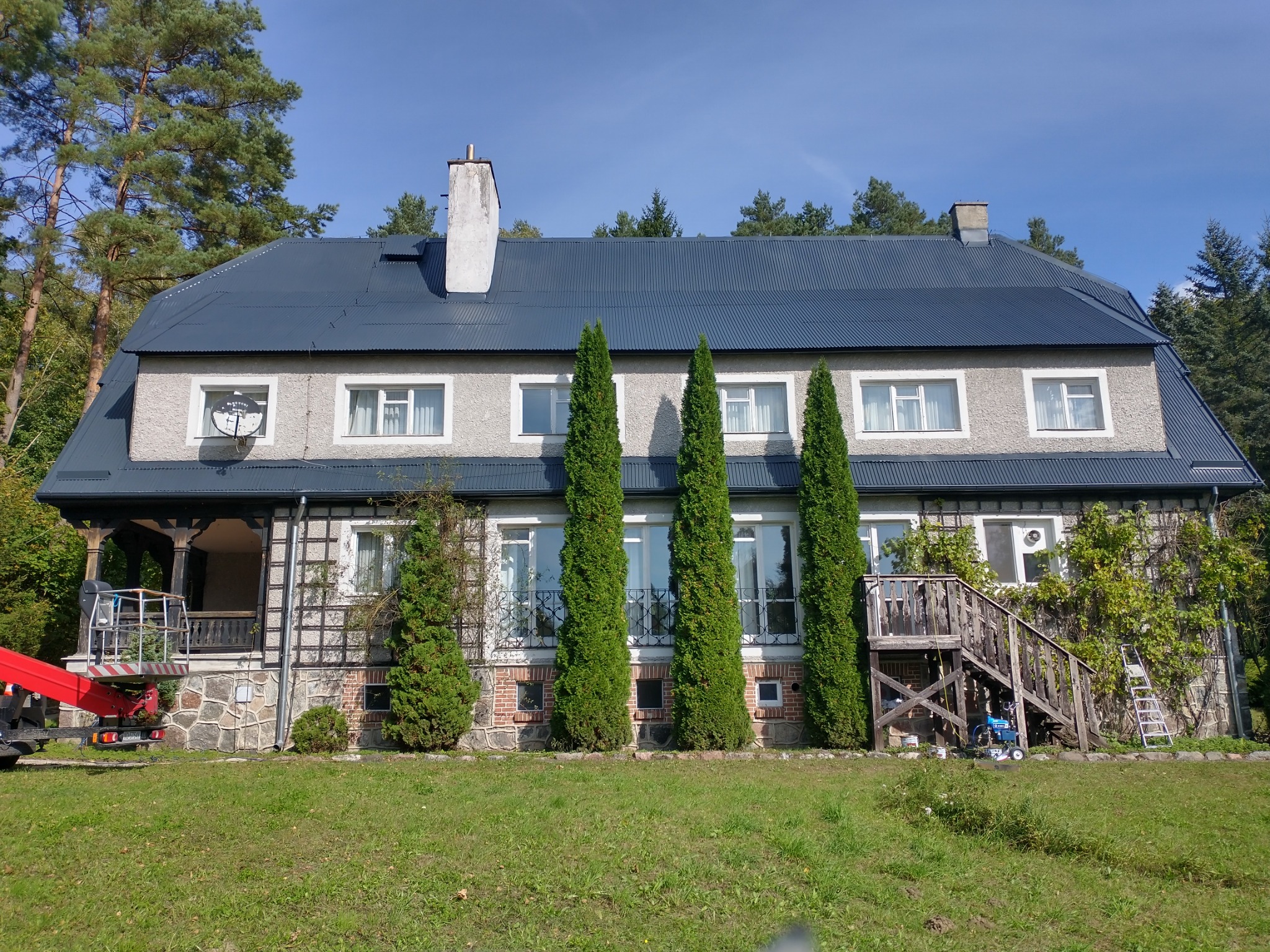 Duży, dwupiętrowy dom z ciemnoszarym dachem po renowacji, widoczny podnośnik koszowy i sprzęt malarski sugerujący świeże malowanie elewacji, otoczony zielenią.