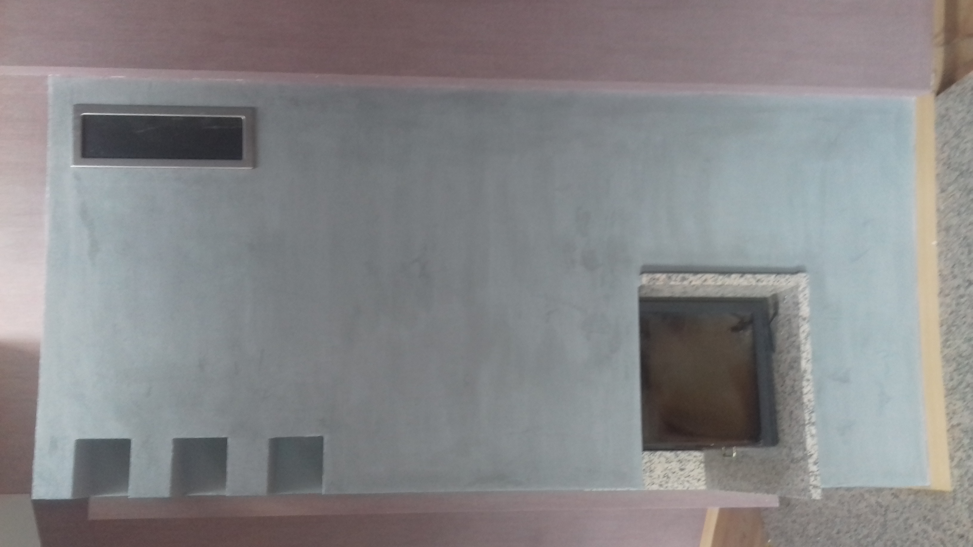 Ściana po szpachlowaniu z otworem na kominek obudowanym płytkami i prostokątnym otworem wentylacyjnym powyżej, widoczne trzy małe kwadratowe wnęki u dołu.