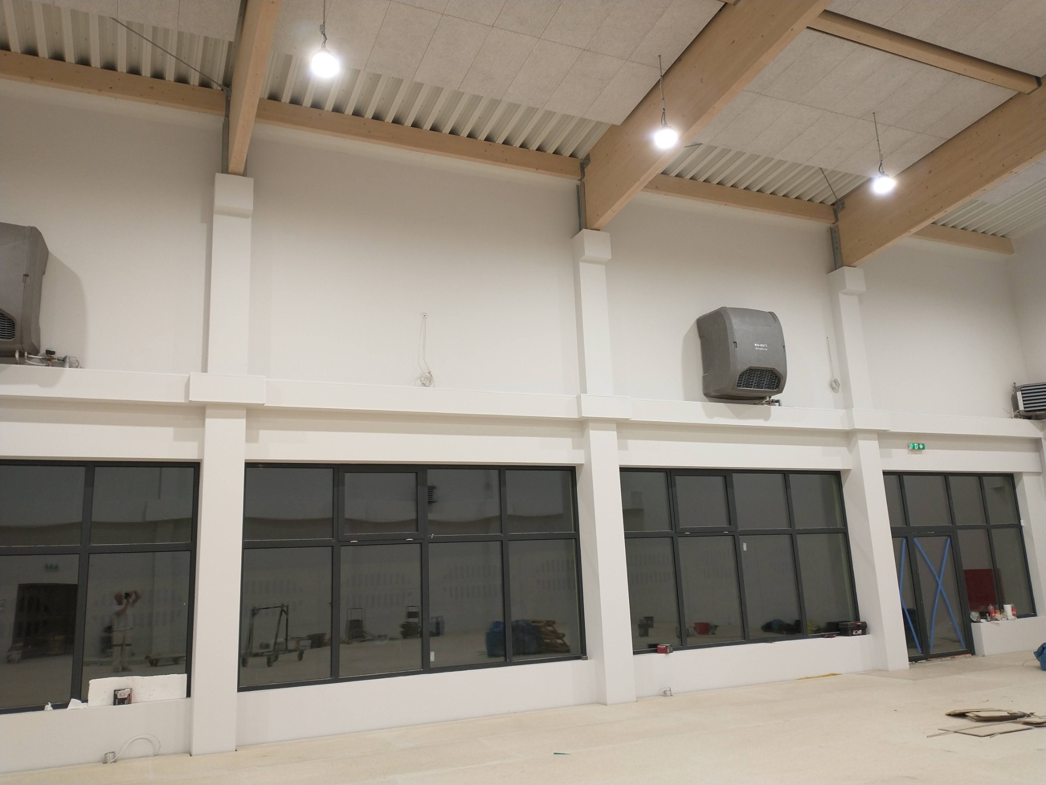 Wnętrze hali z drewnianą konstrukcją dachu, białymi ścianami, dużymi oknami i zamontowanymi klimatyzatorami, widoczny cień osoby robiącej zdjęcie w szybie.