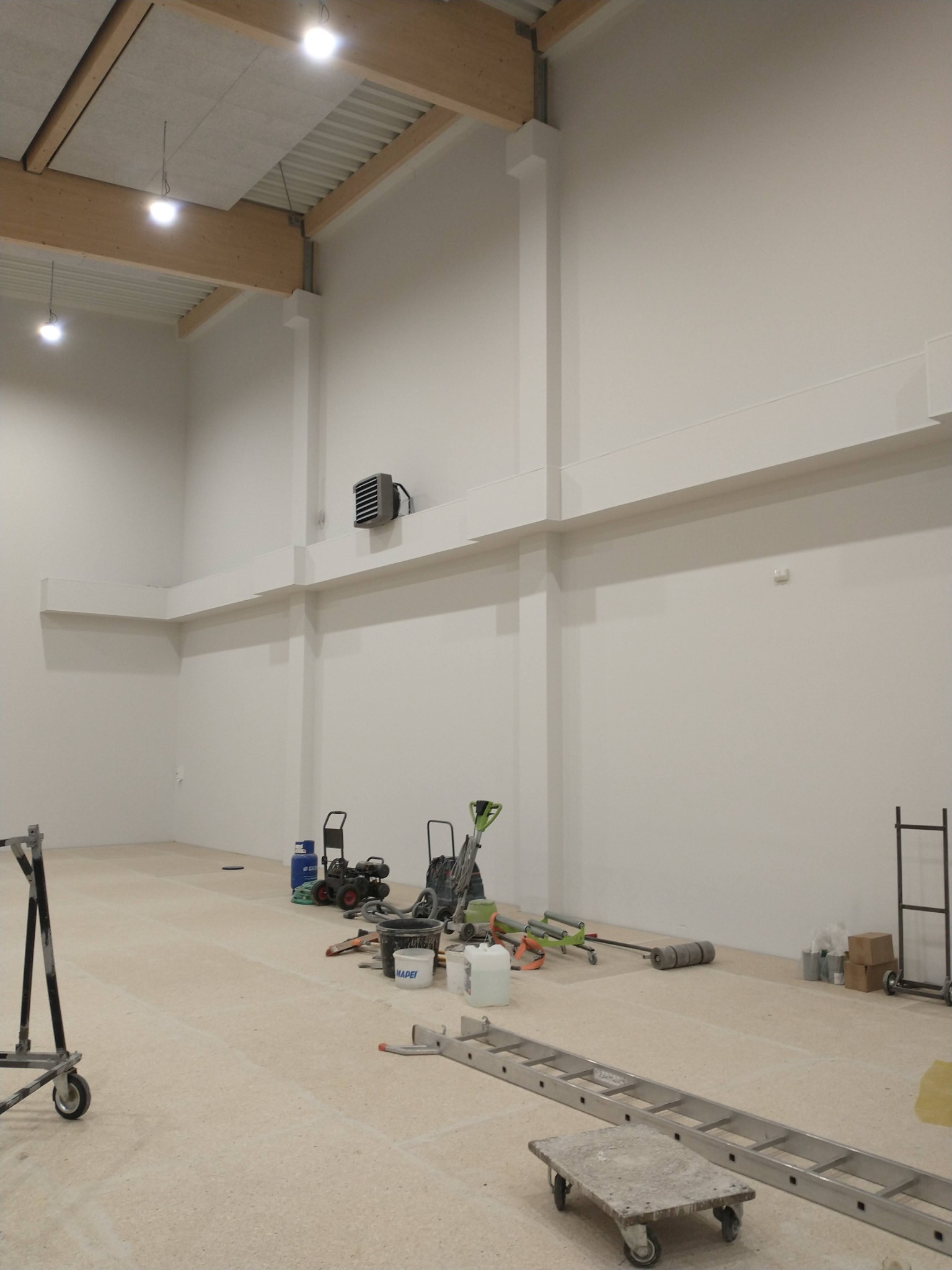 Wnętrze hali przemysłowej z białymi ścianami, drewnianą konstrukcją stropu i sprzętem malarskim na podłodze, w tym agregatami, wałkami, drabiną i pojemnikami z farbą.