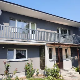 Dwukondygnacyjny dom z szarą elewacją, balkonem z szarą balustradą i anteną satelitarną, otoczony zielenią i kwitnącymi krzewami róż.