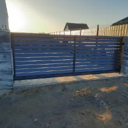 Nowoczesna brama przesuwna w kolorze grafitowym z poziomymi panelami, osadzona na betonowych słupkach z szarymi płytami imitującymi kamień, widok z zewnątrz na tle domu i ogrodzenia.