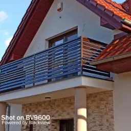 Nowoczesna balustrada balkonowa z poziomych, czarnych profili na tle domu z czerwoną dachówką, widok z dołu.
