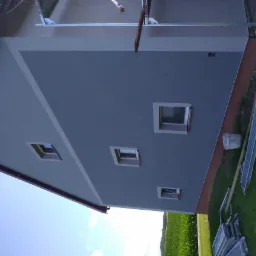 Dwukolorowa elewacja domu z oknami, widok z dołu, z elementami rusztowania leżącymi na trawie.