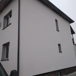 Świeżo otynkowana elewacja domu jednorodzinnego z widocznymi oknami o wąskich ramach i anteną satelitarną, na tle pochmurnego nieba i sąsiedniego budynku.