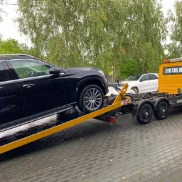 Czarny SUV Mercedes na żółtej platformie holowniczej, widoczny numer telefonu na kabinie lawety, mokra kostka brukowa i drzewa w tle.